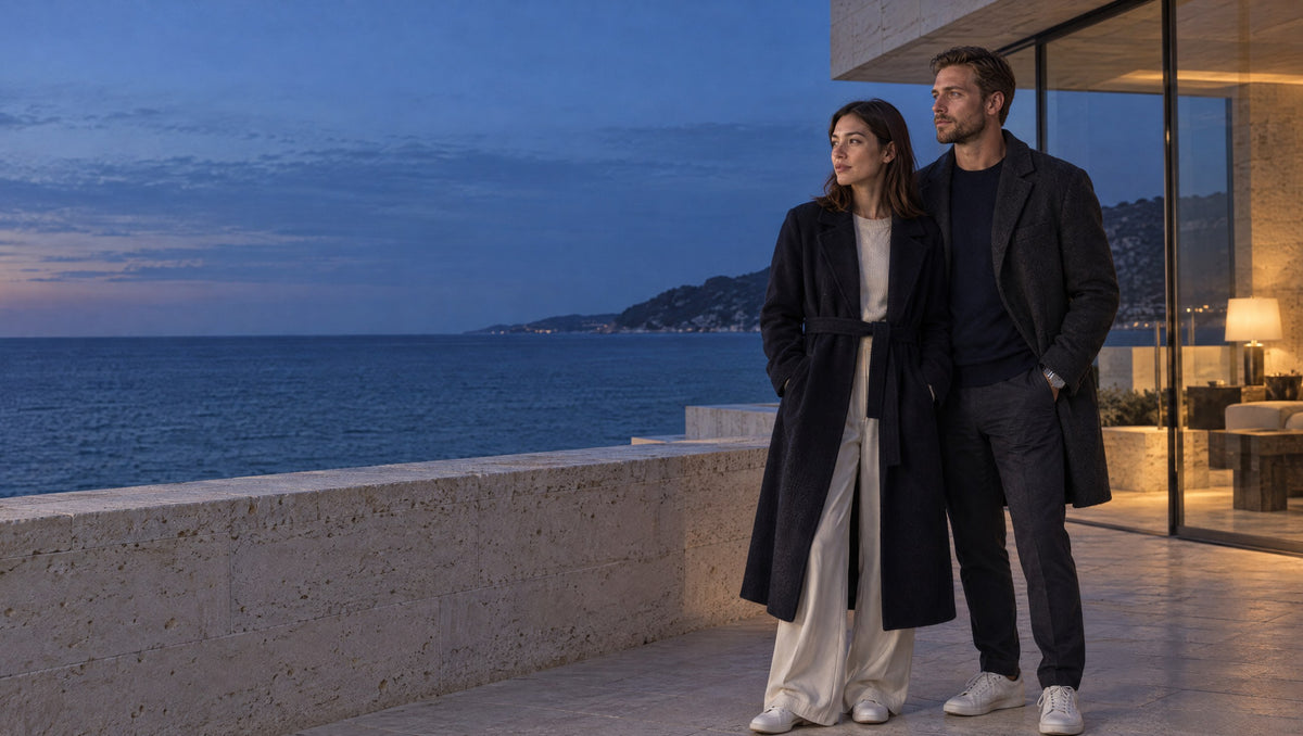 A woman and a man in dark tailored coats stand on a limestone terrace by the sea at blue hour